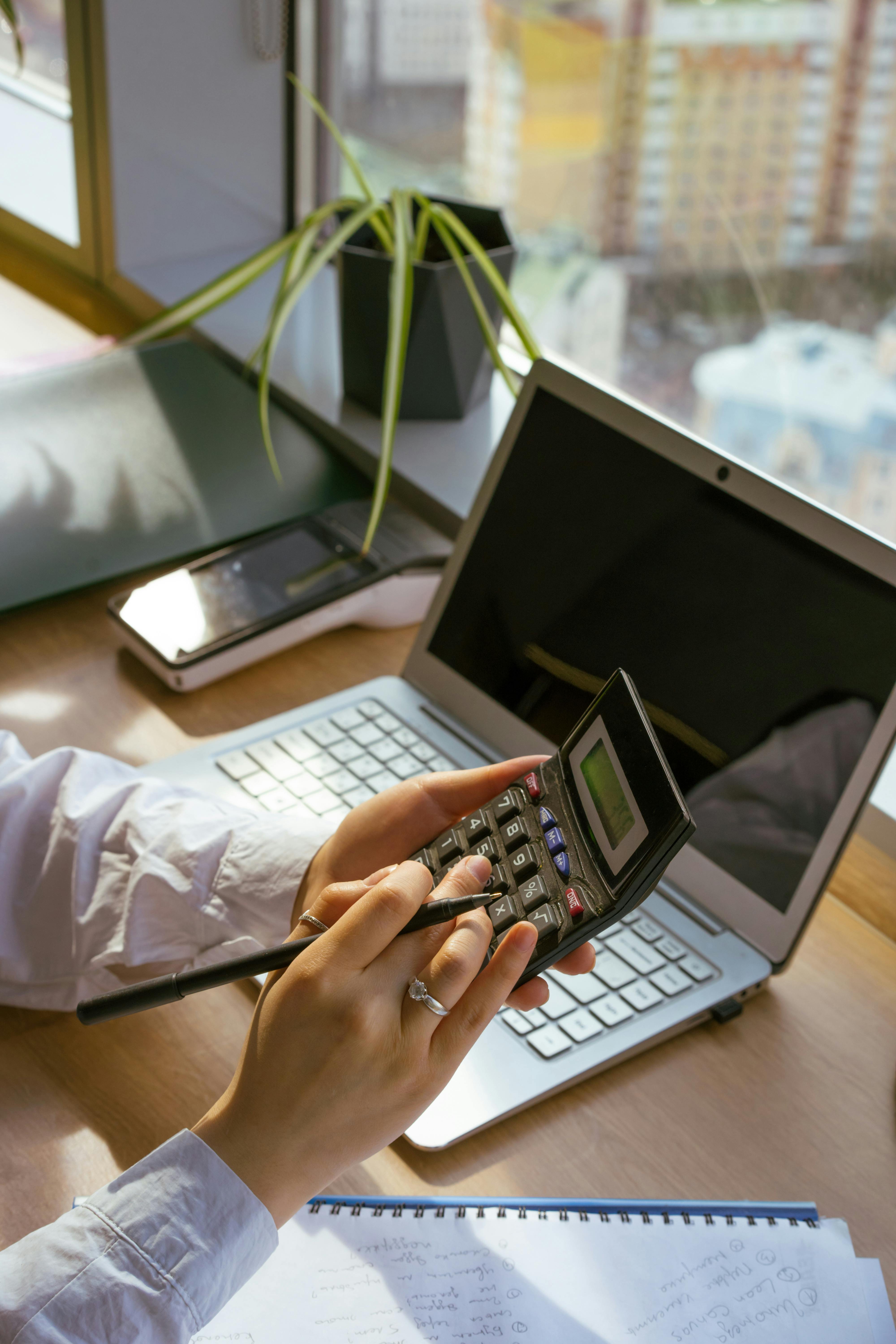 Person using a calculator beside a laptop while reviewing handwritten monthly figures